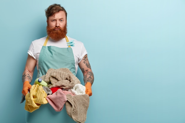 Homem ruivo ocupado e insatisfeito carrega a bacia cheia de roupa para a máquina de lavar, chateado com o trabalho duro e os deveres domésticos