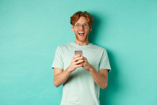Homem ruivo surpreso de óculos, olhando espantado com a câmera depois de ler a oferta promocional no smartphone, de pé contra um fundo turquesa.