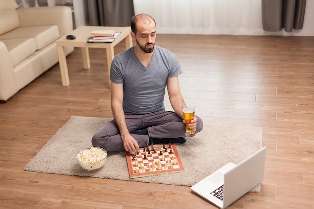 Foto grátis homem segurando o copo de cerveja enquanto joga xadrez online durante o auto-isolamento.