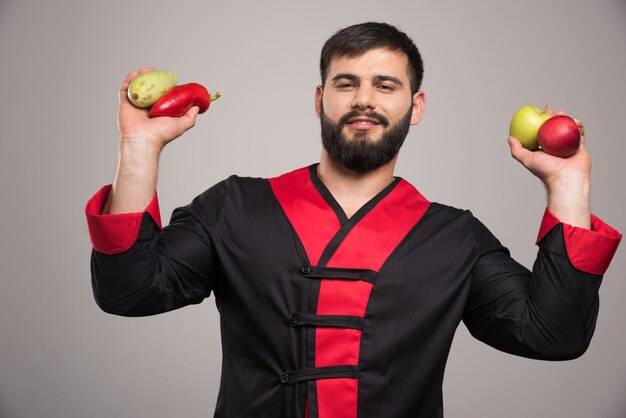 Homem segurando pimenta vermelha, maçãs e abobrinha.