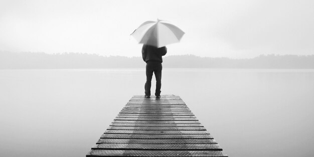 Homem segurando um guarda-chuva em um cais perto de um lago tranquilo.