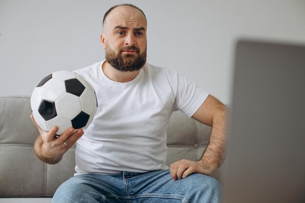 Homem segurando uma bola de futebol e assistindo futebol online em casa
