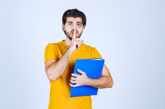 Homem segurando uma pasta azul e pedindo silêncio.