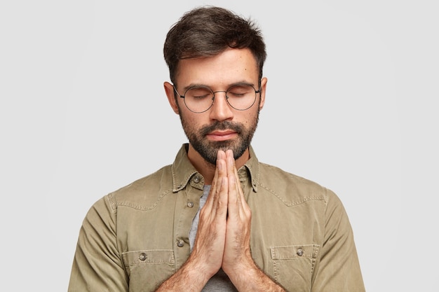 Foto grátis homem sem barbear concentrado fica em gesto de oração, mantém as palmas das mãos pressionadas