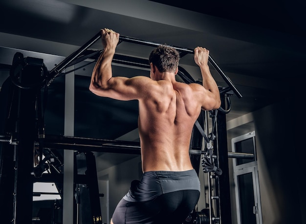 Homem sem camisa atlético fazendo pull ups na barra horizontal em um clube de ginástica.