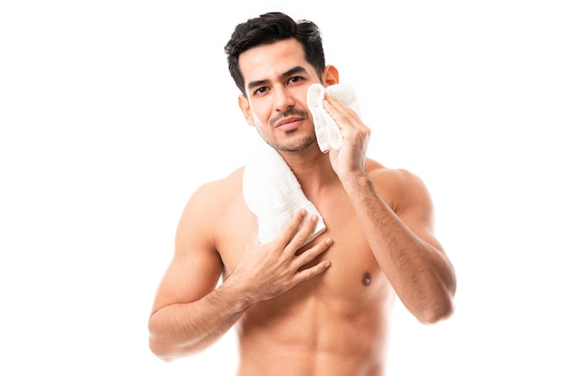 Homem sem camisa bonito, limpando o rosto usando uma toalha em pé contra um fundo branco