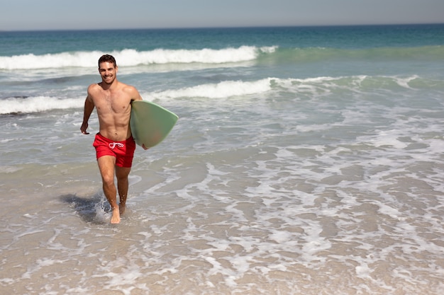 Homem sem camisa com prancha andando na praia ao sol