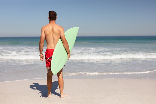 Homem sem camisa com prancha de surf em pé na praia ao sol