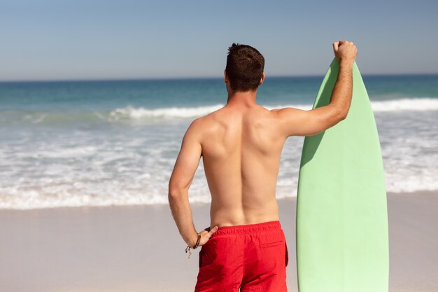 Homem sem camisa com prancha de surf em pé na praia ao sol