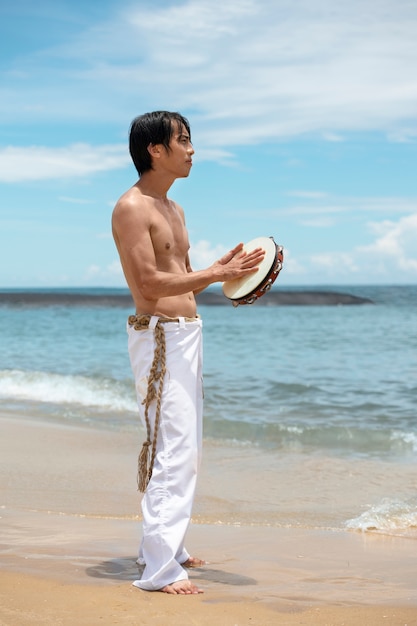 Homem sem camisa praticando capoeira na praia com pandeiro