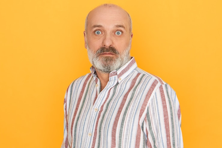 Foto grátis homem sênior careca com a barba por fazer, espantado, vestindo uma camisa listrada, arregalando os olhos e sendo pego de surpresa, recebendo uma notícia inesperada posando isolada contra o fundo amarelo da parede do estúdio