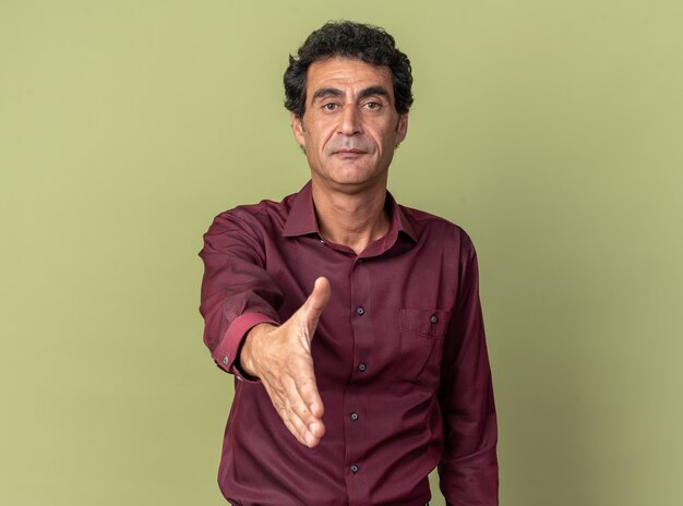 Homem sênior com camisa roxa e gesto de saudação com o braço, parecendo confiante em pé sobre um fundo verde