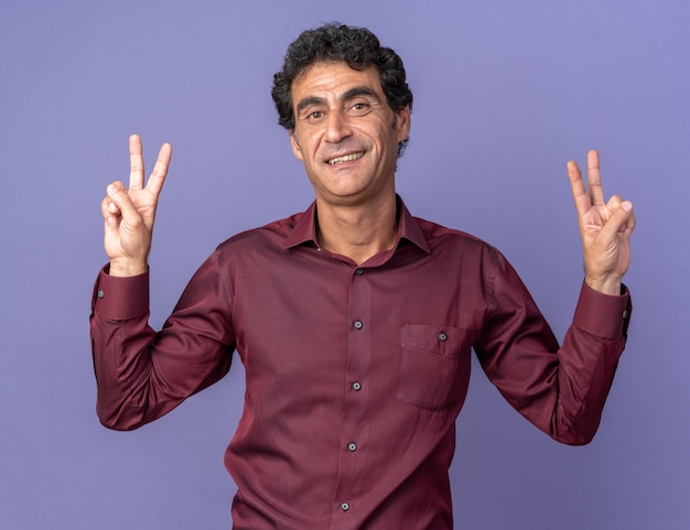 Homem sênior com camisa roxa olhando para a câmera feliz e positivo mostrando o sinal de "V" em pé sobre o azul
