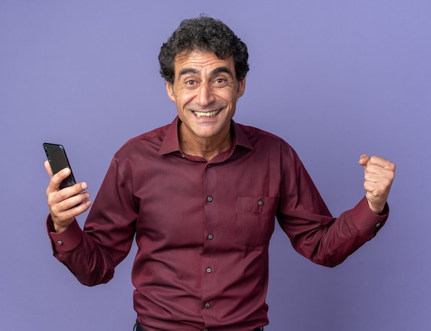 Homem sênior com camisa roxa segurando o punho cerrado de smartphone feliz e animado