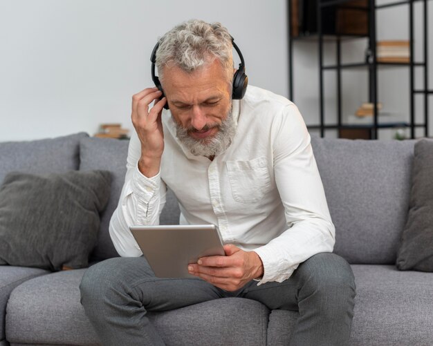 Homem sênior em casa no sofá usando tablet e fones de ouvido