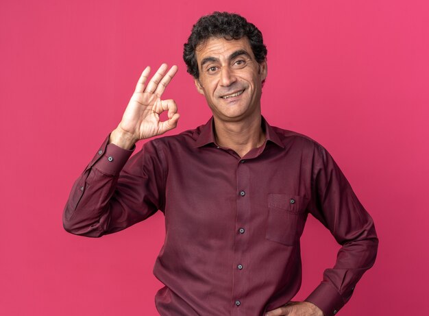 Homem sênior feliz e positivo de camisa roxa, olhando para a câmera, sorrindo alegremente, fazendo sinal de ok em pé sobre o fundo rosa