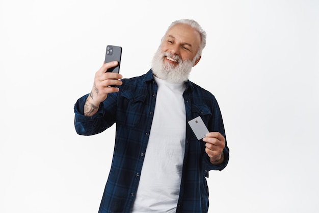 Homem sênior feliz tirando uma selfie com seu cartão de crédito, sorrindo como pagando online com identificação de rosto no aplicativo do smartphone, fazendo compras em uma loja de internet, encostado na parede branca