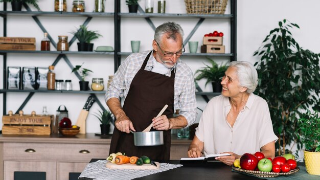 Homem sênior, preparando alimento, olhar, livro, segure, por, mulher sênior