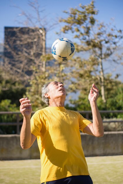 Homem sênior satisfeito com bola de futebol. Homem caucasiano com cabelo grisalho curto em roupas esportivas jogando bola no campo de esporte sozinho. Futebol, esporte, conceito de lazer