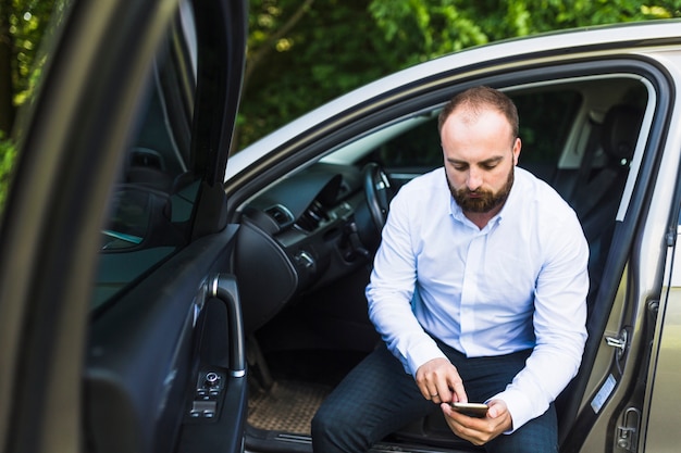 Homem sentado em um carro com a porta aberta, olhando para a tela do telefone móvel