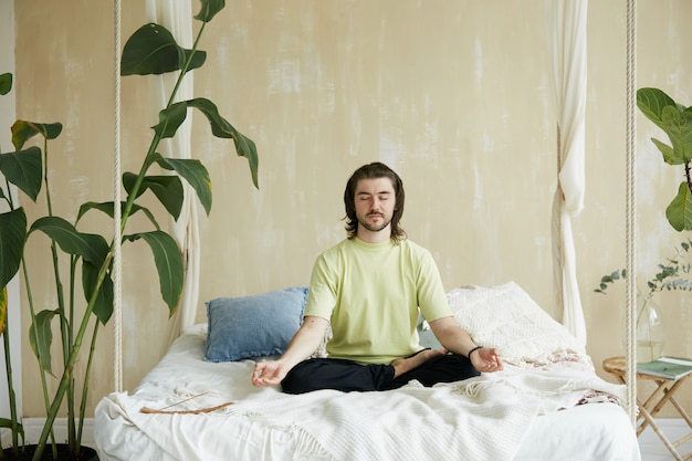 Homem sentado na cama com os olhos fechados e meditando, belo meditador se concentrando em casa com um bastão de aroma na cama