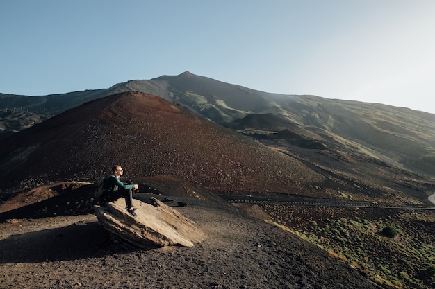 Homem sentado na rocha e apreciando a bela paisagem do vulcão Etna, na Sicília