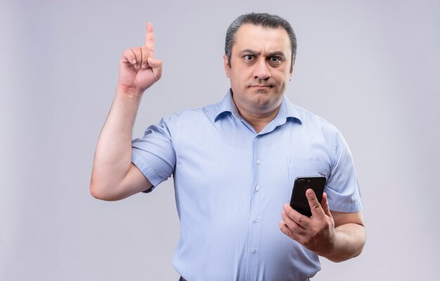 Homem sério de meia-idade usando camisa listrada azul, proibindo algo levantando o dedo indicador e segurando o telefone celular com a outra mão em um fundo branco