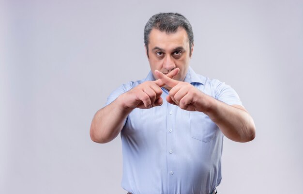 Homem sério de meia-idade vestindo camisa azul, mostrando a forma do sinal x com os dedos em um fundo branco