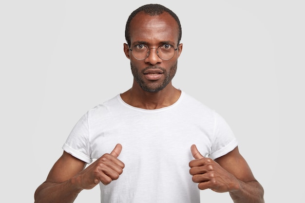 Homem sério e rabugento aponta para si mesmo, surpreso por ter muitos deveres, usa uma camiseta branca casual, isolado na parede do estúdio