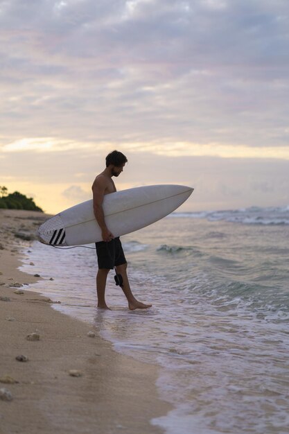 Homem sexy surfista com prancha de surf. Bonito jovem atleta masculino segurando a prancha de surf com cabelo molhado nas férias de esporte de praia de verão. Destino de viagem esportiva. Estilo de vida surf.