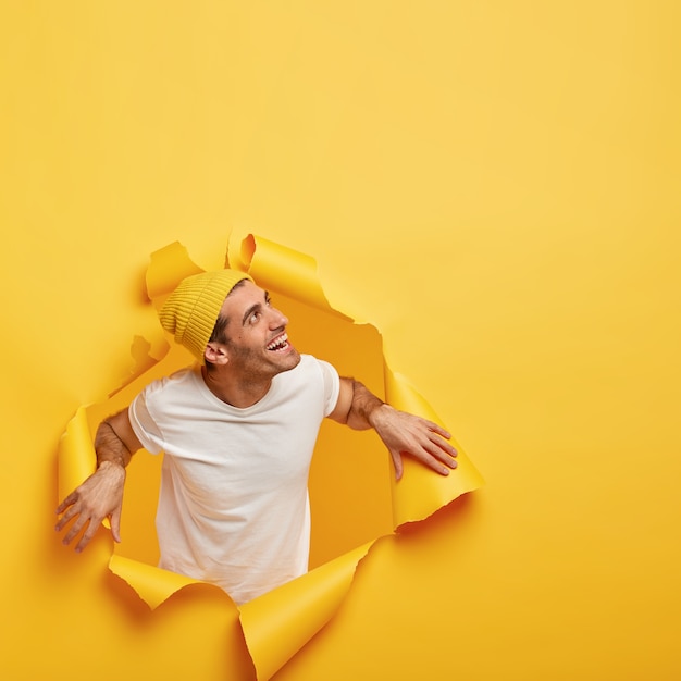 Homem simpático e satisfeito desvia o olhar, em pé em um buraco de papel rasgado, vestido com uma camiseta branca