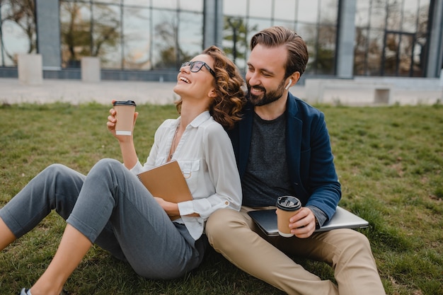 Homem sorridente atraente e mulher conversando sentado na grama em um parque urbano, fazendo anotações