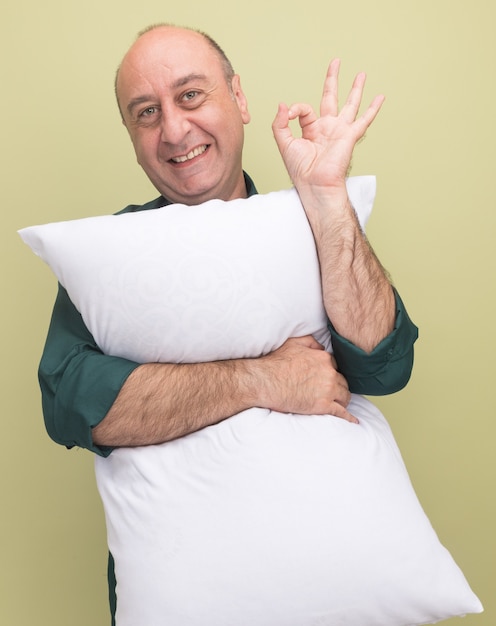 Homem sorridente de meia-idade vestindo uma camiseta verde abraçou o travesseiro mostrando um gesto de aprovação isolado na parede verde oliva