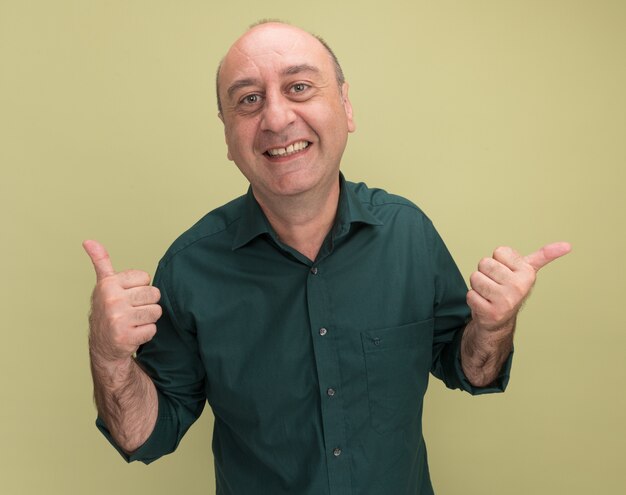 Homem sorridente de meia-idade vestindo uma camiseta verde mostrando o polegar para cima e apontando para o lado isolado na parede verde oliva com espaço de cópia