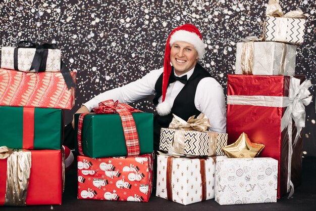 Homem sorridente de terno com chapéu de papai noel posando com um presente de natal rodeado de neve