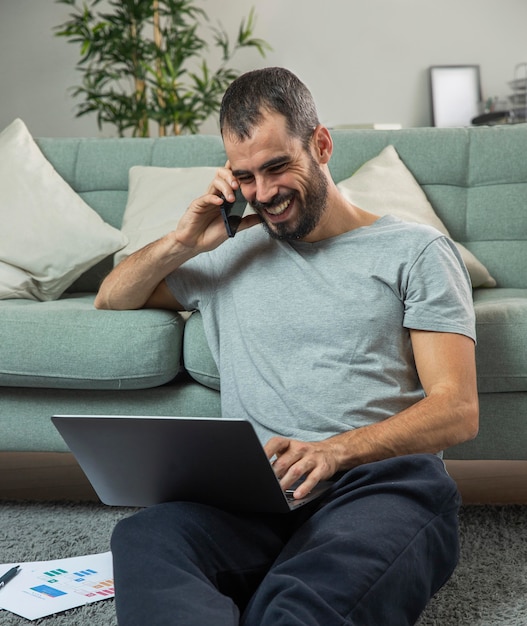 Foto grátis homem sorridente falando ao telefone em casa enquanto trabalha no laptop