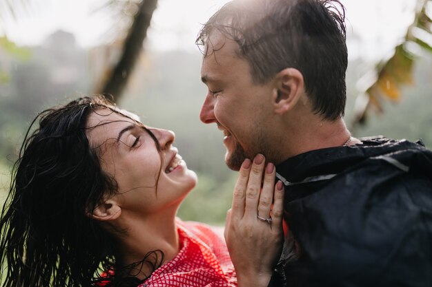 Homem sorridente na capa de chuva, olhando com amor para uma mulher morena. Rindo casal romântico dançando na natureza.