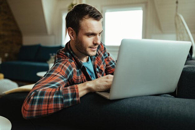 Homem sorridente navegando na net no laptop enquanto relaxa no sofá em casa