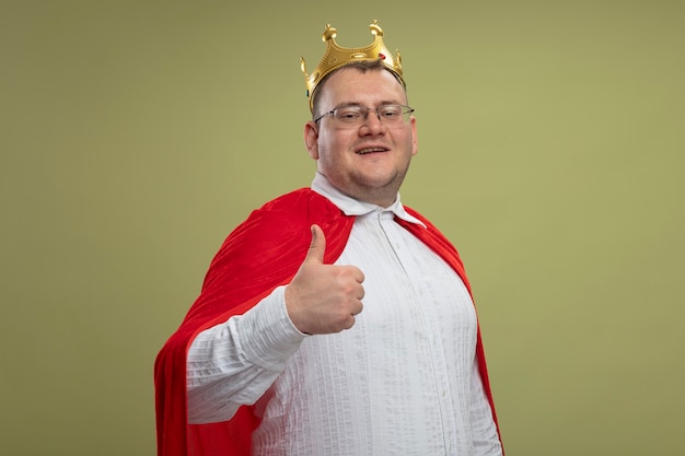 Homem sorridente super-herói eslavo adulto com capa vermelha usando óculos e coroa, olhando para a câmera, mostrando o polegar isolado em um fundo verde oliva com espaço de cópia