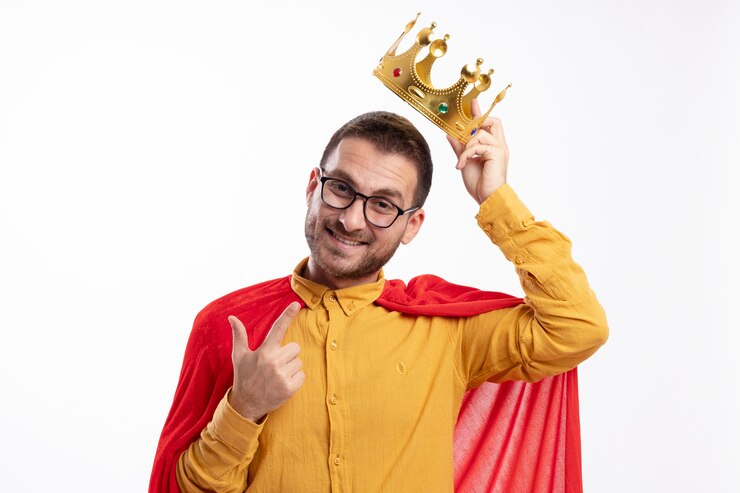 Foto grátis homem super-herói sorridente com óculos ópticos e capa vermelha segurando a coroa sobre a cabeça e apontando para a frente, isolada na parede branca