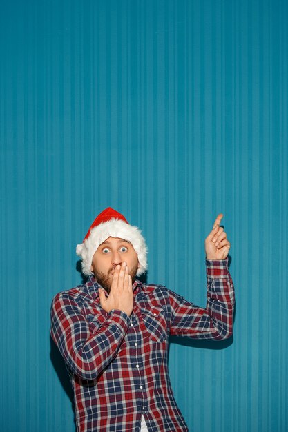 Homem surpreso com chapéu de Papai Noel mostrando qualquer coisa sobre fundo azul