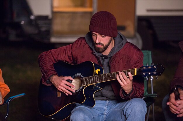 Homem tocando violão em uma noite fria de outono em um acampamento para seus amigos com uma van de campista retrô ao fundo.