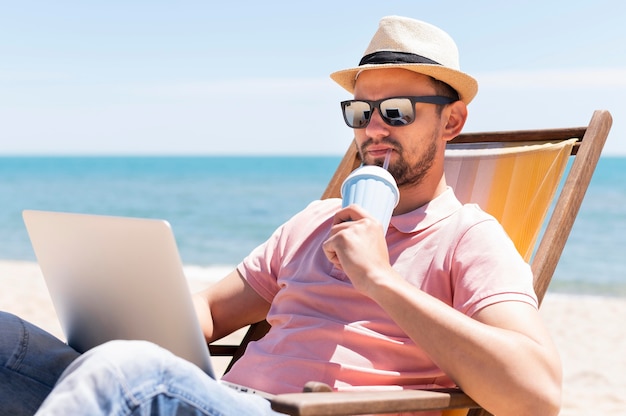 Homem tomando uma bebida na praia e trabalhando no laptop