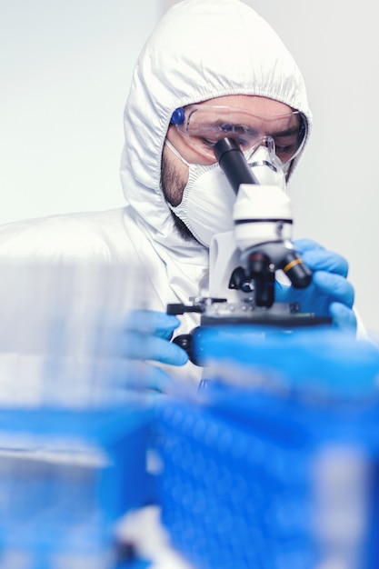 Homem trabalhador em traje de ppe usando microscópio fazendo pesquisa de coronavírus Cientista em traje de proteção sentado no local de trabalho usando tecnologia médica moderna durante a epidemia global.