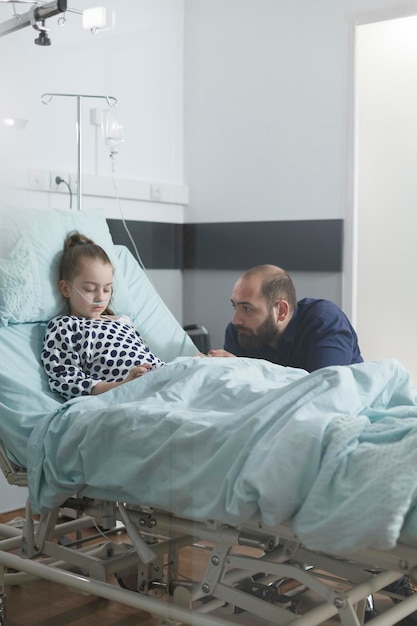 Foto grátis homem triste e ansioso assistindo filha doente dormindo na cama da clínica enquanto na enfermaria de pacientes do hospital pediátrico. em tratamento sedado criança doente descansando enquanto pai atento cuidando dela.