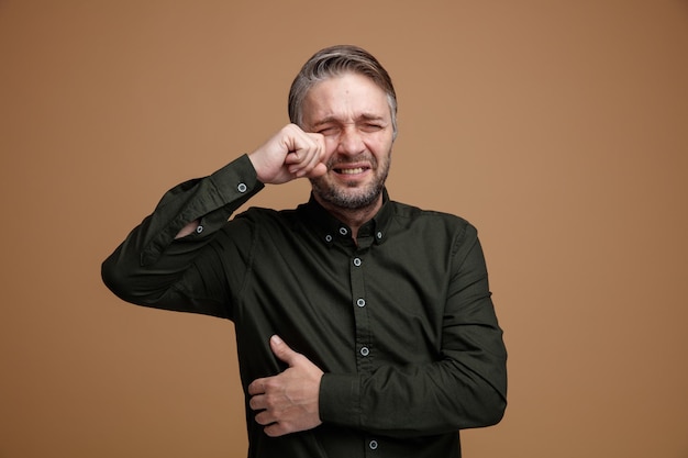 Homem triste triste de meia-idade com cabelos grisalhos na camisa de cor escura chorando esfregando o olho com a mão em pé sobre fundo marrom