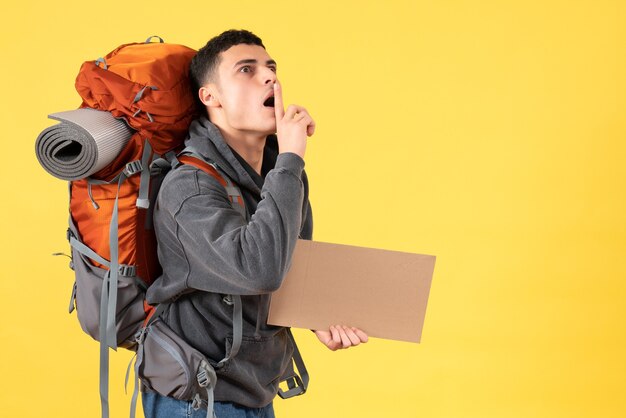 Homem viajante com mochila segurando um papelão e fazendo sinal de silêncio