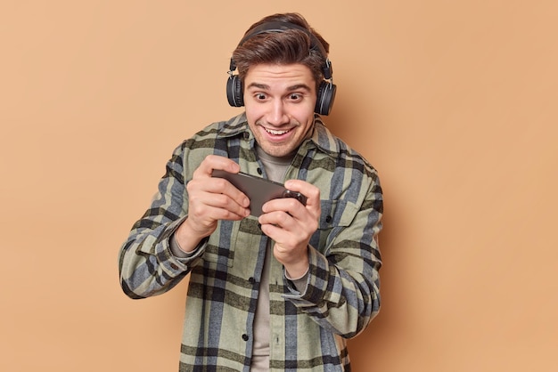 Homem viciado joga videogame segura smartphone horizontalmente tenta passar nível difícil usa fones de ouvido estéreo nas orelhas vestida com camisa quadriculada isolada sobre fundo bege tecnologia