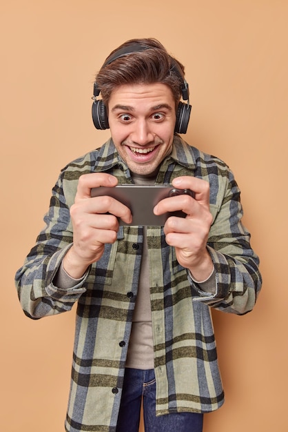 Homem viciado otimista joga videogame no smartphone e olha para a tela do celular fica contente de usar fones de ouvido sem fio nas orelhas vestido com uma camisa xadrez isolada sobre um fundo bege