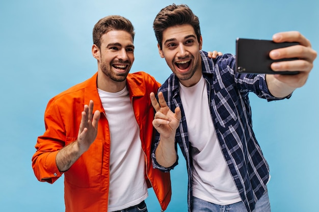 Homens alegres e encantadores tiram selfie de bom humor em fundo azul Homem legal de jaqueta laranja acena com a mão em saudação Cara de camisa quadriculada segura telefone e mostra sinal de paz